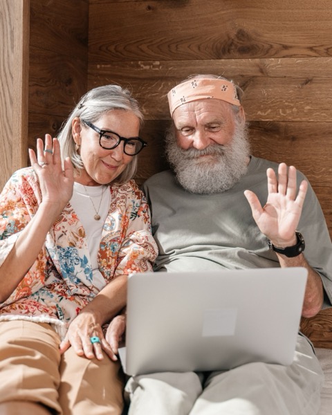 An older couple waving at a laptop from their sofa